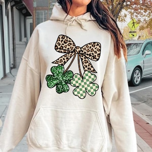 May include: Sand-colored hoodie featuring a St. Patrick's Day design. The design includes a leopard print bow and two shamrocks, one green glitter and one green and white checkered. The hoodie has a front pocket and a drawstring hood.