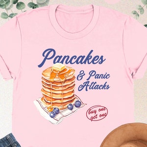 May include: Pink t-shirt with a graphic of a stack of pancakes with syrup and butter, and the text "Pancakes & Panic Attacks" and "buy one get one".