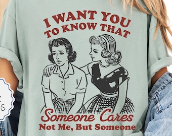 May include: A light green t-shirt with the text "I WANT YOU TO KNOW THAT" above a black and white illustration of two women. The text "Someone Cares Not Me, But Someone" is below the illustration. The shirt also has a "MORE COLOR OPTIONS" button.