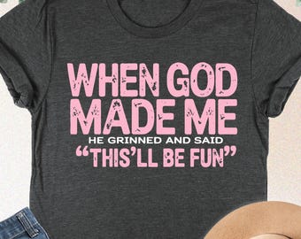 May include: Dark grey heather t-shirt with the text "WHEN GOD MADE ME HE GRINNED AND SAID 'THIS'LL BE FUN'" in pink. The image also shows blue jeans and a tan hat.