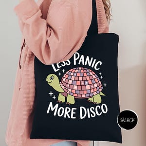 May include: Black tote bag featuring a cartoon turtle with a disco ball shell. The text on the bag reads "LESS PANIC MORE DISCO" in white. The turtle is green and the disco ball is pink, purple, and white.