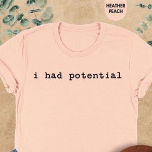 May include: A heather peach short-sleeve t-shirt with the text i had potential printed in black typewriter-style font across the chest. The shirt is paired with blue denim jeans and a brown wide-brimmed hat, creating a casual ensemble.