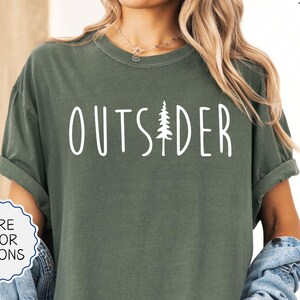 May include: A green t-shirt with the text "OUTSIDER" in white lettering and a small pine tree graphic. The shirt is shown on a model wearing denim shorts and a denim jacket.