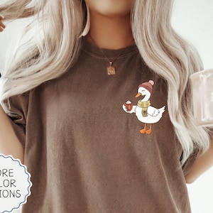 May include: An espresso-colored t-shirt featuring a graphic of a white goose wearing a pink beanie and a yellow scarf while holding a small brown mug. The shirt has a crew neckline and short sleeves, with a simple, clean design on the chest area.