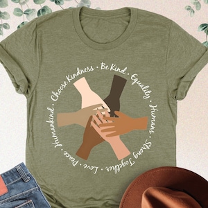 May include: A green t-shirt with a circle design featuring five hands of different skin tones clasped together. The text around the circle reads "Choose Kindness. Be Kind. Equality. Humans. Strong Together. Love. Peace. Humankind."