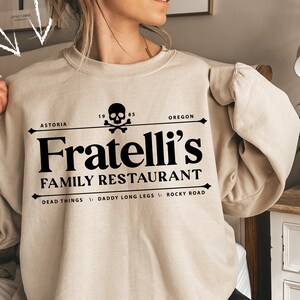 May include: A beige sweatshirt with a black graphic print of a skull and crossbones, the text "Fratelli's Family Restaurant", and the words "Astoria", "Oregon", "Dead Things", "Daddy Long Legs", and "Rocky Road".