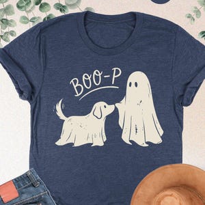 May include: A heather navy t-shirt featuring a cream-colored graphic of a dog and a ghost wearing sheets, with the text "BOO-P" above them. The shirt is displayed alongside a pair of blue denim jeans and a tan wide-brimmed hat.