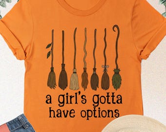 May include: An orange short-sleeved t-shirt featuring a graphic of eight different broomsticks arranged in a row above the black text that reads, a girl's gotta have options. The shirt is paired with blue denim jeans and a beige fedora hat.
