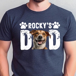 May include: A navy blue t-shirt with a white graphic of a dog's head and the text "ROCKY'S DAD" in a distressed font.