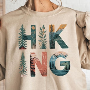 Pode incluir: Uma camisa de moletom bege com a palavra "HIKING" impressa na frente. As letras s&atilde;o compostas por ilustra&ccedil;&otilde;es de montanhas, &aacute;rvores e outras cenas da natureza.