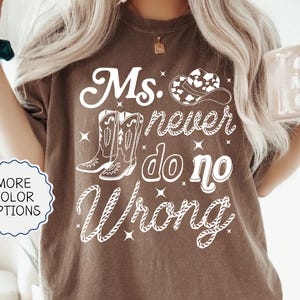 May include: Espresso-colored t-shirt with the text "Ms. Never Do No Wrong" in white script. The design includes cowboy boots and a hat. A "More Color Options" button is visible.
