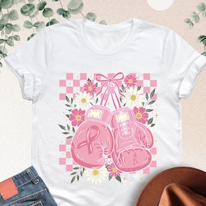 May include: White t-shirt featuring a pink boxing glove design with a pink ribbon, flowers, and a checkered background. The shirt is paired with blue jeans and a brown hat.