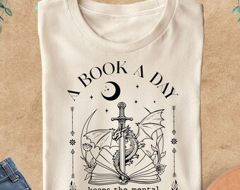May include: Cream-colored t-shirt with a black graphic featuring a dragon, sword, and open book, with the text "A BOOK A DAY keeps the mental breakdown away." The shirt is displayed with blue jeans and a brown hat.