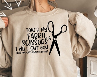 May include: Beige sweatshirt with the text "TOUCH MY FABRIC & SCISSORS I WILL CUT YOU (but not with these scissors)" and a black scissors graphic. The sweatshirt is a neutral color, suitable for various styles.