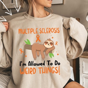 May include: A beige sweatshirt with the text "MULTIPLE SCLEROSIS" and a cartoon sloth with an orange ribbon. The text below the sloth reads "I'm Allowed To Do WEIRD THINGS!"