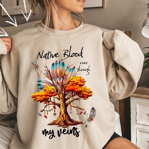 May include: A beige sweatshirt with a graphic of a tree with feathers and the text "Native Blood runs through my veins".