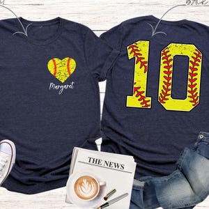 K&ouml;nnte beinhalten: Zwei marineblaue T-Shirts mit Softball-Motiv. Das vordere Shirt hat ein gelbes Softball-Herz mit roten N&auml;hten und den Namen "Margaret" darunter gedruckt. Das hintere Shirt hat eine gro&szlig;e gelbe Nummer "10" mit roten N&auml;hten, die einem Softball &auml;hnelt.