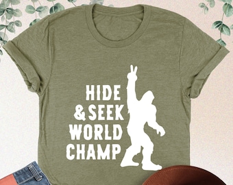 May include: A green t-shirt with a white silhouette of a Bigfoot raising its hand in a peace sign. The text "HIDE & SEEK WORLD CHAMP" is printed on the shirt.