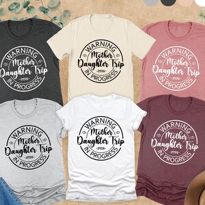 K&ouml;nnte beinhalten: Eine Auswahl an kurz&auml;rmeligen T-Shirts in verschiedenen Farben, darunter Grau, Wei&szlig; und Burgunderrot. Jedes Shirt zeigt eine runde Grafik mit dem Text "WARNING Mother Daughter Trip -2026- IN PROGRESS".