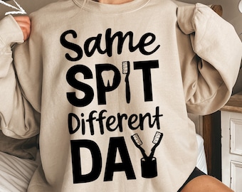 May include: A beige sweatshirt with the text "Same Spit Different Day" in black. The text is styled with a tooth and toothbrush graphic.