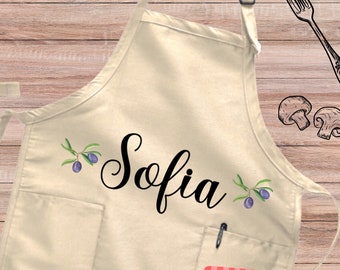 May include: A beige kitchen apron with a large pocket and a black cursive name "Sofia" with olive branch designs. The apron has adjustable neck straps and a tie at the waist.