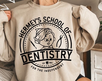 Op de afbeelding: Een beige sweatshirt met een zwart grafisch ontwerp met de tekst "Hermey's School of Dentistry" met een cartoonfiguur dat een tandheelkundig gereedschap vasthoudt. De grafiek bevindt zich in een cirkel met een gestippelde rand en de tekst "Est. 1964" en "For the Independent" onder de cirkel.