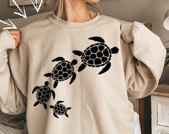 May include: A beige sweatshirt with a black silhouette of four sea turtles of different sizes. The turtles are arranged in a line, with the largest turtle at the top and the smallest turtle at the bottom.