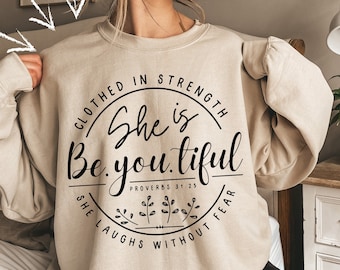 Może przedstawiać: Beżowa bluza z czarnym, okrągłym wzorem. Wzór zawiera słowa "CLOTHED IN STRENGTH", "She is Beautiful" i "SHE LAUGHS WITHOUT FEAR". Tekst "PROVERBS 31:25" jest również zawarty.