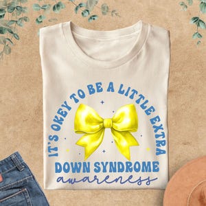 May include: Cream-colored t-shirt with a yellow bow graphic and the text "IT'S OKEY TO BE A LITTLE EXTRA DOWN SYNDROME awareness" in blue. The shirt is folded and displayed with blue jeans and a brown hat.