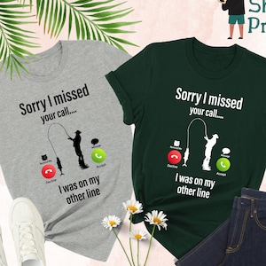 May include: Two t-shirts, one gray and one forest green, with a fishing-themed graphic. The text on the shirts reads "Sorry I missed your call... I was on my other line." White sneakers and a pair of dark blue jeans are also visible.