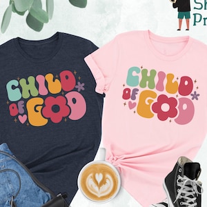 Op de afbeelding: Twee T-shirts met de tekst "Child of God" in een retro, kleurrijk lettertype. De shirts zijn in heideblauw en roze.
