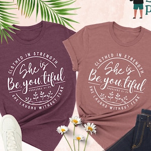 May include: Two heather t-shirts, one maroon and one mauve, with the text "She is Be.you.tiful" in a circular design. The shirts are paired with white sneakers and dark blue jeans. The words "Fast Shirt Print" are also visible.