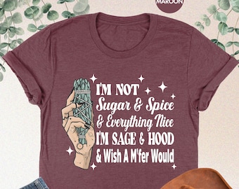 Op de afbeelding: Een heide-marron t-shirt met een wit grafisch ontwerp met een hand die een salie-stokje vasthoudt. De tekst op het shirt luidt "I'm not sugar & spice & everything nice I'm sage & hood & wish a m'fer would".
