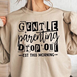 May include: A sand-colored crewneck sweatshirt featuring black text that reads GENTLE parenting DROP OUT EST THIS MORNING. The design includes stylized lettering and small star accents centered on the front of the garment.