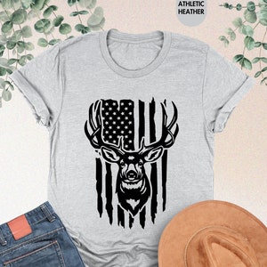 Puede incluir: Una camiseta gris con una silueta negra de una cabeza de ciervo delante de una bandera estadounidense.