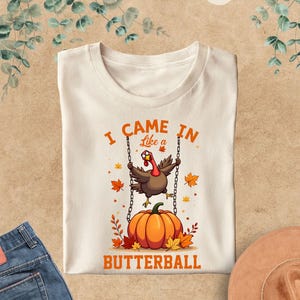 以下が含まれることがあります： クリーム色のTシャツ。カボチャの上でブランコに乗る七面鳥のイラストと「I CAME IN Like a BUTTERBALL」の文字。秋の葉がカボチャを囲んでいます。シャツは茶色の表面に折りたたまれています。