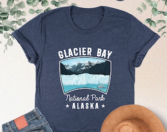 Peut inclure: Un t-shirt bleu marine chiné avec un motif représentant le parc national de Glacier Bay en Alaska. Le motif représente une chaîne de montagnes, un glacier et un bateau sur une étendue d'eau. Le texte "GLACIER BAY", "National Park" et "* ALASKA *" est imprimé en blanc sur le t-shirt.