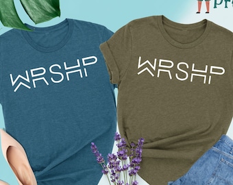 May include: Two t-shirts with the word "WRSHP" printed in white on a teal and olive green background. The shirts are laid out on a light blue surface with a bouquet of lavender flowers in the center.