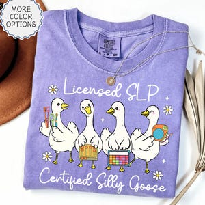 May include: A violet Comfort Colors t-shirt with a playful design of four white geese, each holding speech therapy tools. The text reads "Licensed SLP Certified Silly Goose". A gold necklace is draped on the shirt.