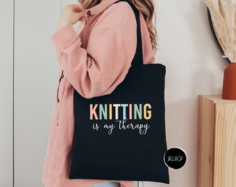 May include: Black tote bag with the text "Knitting is my therapy" in colorful letters. The bag has a black strap and is being held by a person.