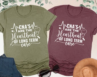 May include: Two t-shirts with the text "CNA's are the Heartbeat of Long Term Care" in white on a maroon and a green shirt. The shirts are laid out on a surface with a pair of jeans and sunglasses.
