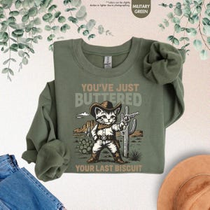 May include: Military green sweatshirt featuring a cartoon cat in a cowboy hat, holding a gun, with the text "YOU'VE JUST BUTTERED YOUR LAST BISCUIT". The design includes a desert scene with cacti and mountains.