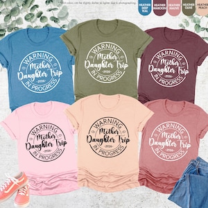 K&ouml;nnte beinhalten: Eine Auswahl an T-Shirts in verschiedenen Farben, darunter T&uuml;rkis, Olive und Rosa, mit dem Text "WARNING Mother Daughter Trip IN PROGRESS -2026-" in einem kreisf&ouml;rmigen Design. Die Shirts werden mit Jeans und Turnschuhen pr&auml;sentiert.