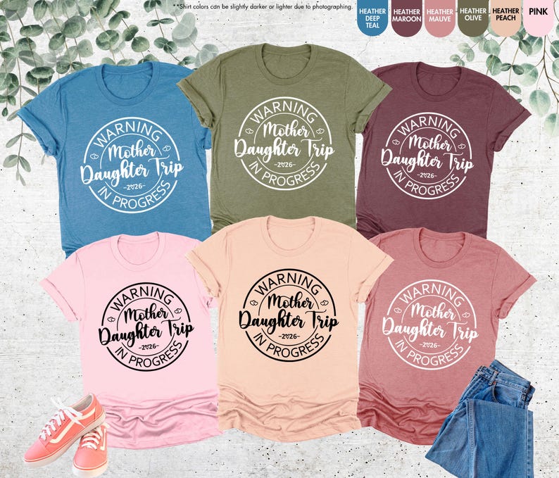 K&ouml;nnte beinhalten: Eine Auswahl an T-Shirts in verschiedenen Farben, darunter T&uuml;rkis, Olive und Rosa, mit dem Text "WARNING Mother Daughter Trip IN PROGRESS -2026-" in einem kreisf&ouml;rmigen Design. Die Shirts werden mit Jeans und Turnschuhen pr&auml;sentiert.
