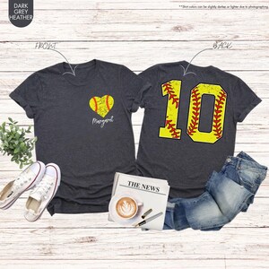 K&ouml;nnte beinhalten: Ein dunkel-heidesgraues T-Shirt mit einem Softball-Herz-Design und dem Namen "Margaret" auf der Vorderseite. Die R&uuml;ckseite des Shirts hat einen gelben Softball mit roten N&auml;hten und die Nummer "10" darauf.