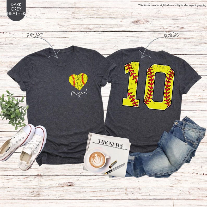K&ouml;nnte beinhalten: Ein dunkel-heidesgraues T-Shirt mit einem Softball-Herz-Design und dem Namen "Margaret" auf der Vorderseite. Die R&uuml;ckseite des Shirts hat einen gelben Softball mit roten N&auml;hten und die Nummer "10" darauf.