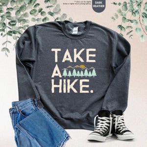 K&ouml;nnte beinhalten: Dunkelgraues Sweatshirt mit dem Text "TAKE A HIKE" in Wei&szlig;, mit einem Berg- und Baum-Motiv. Das Sweatshirt wird mit blauen Jeans und schwarz-wei&szlig;en Turnschuhen kombiniert.