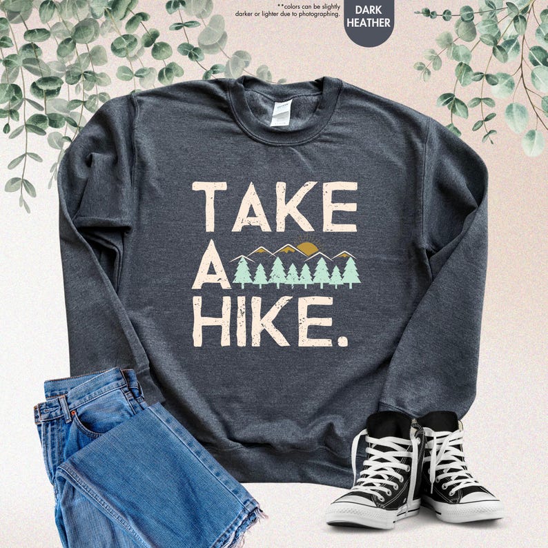 K&ouml;nnte beinhalten: Dunkelgraues Sweatshirt mit dem Text "TAKE A HIKE" in Wei&szlig;, mit einem Berg- und Baum-Motiv. Das Sweatshirt wird mit blauen Jeans und schwarz-wei&szlig;en Turnschuhen kombiniert.