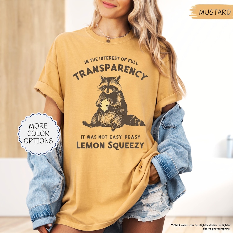 May include: Mustard-colored t-shirt with a raccoon graphic holding a lemon. The text reads "IN THE INTEREST OF FULL TRANSPARENCY, IT WAS NOT EASY PEASY LEMON SQUEEZY." The shirt has rolled-up sleeves and a relaxed fit. "More color options" is also visible.