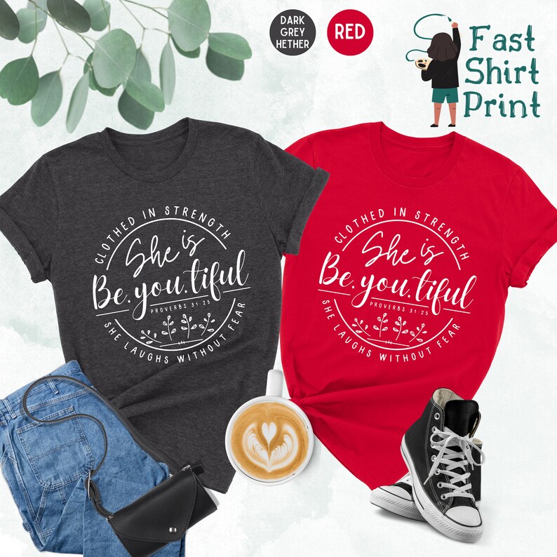 May include: Two t-shirts, one dark gray and one red, each with a white circular design. The design includes the text "Clothed in Strength, She is Beautiful, She Laughs Without Fear." The dark gray shirt is paired with jeans and a black purse.